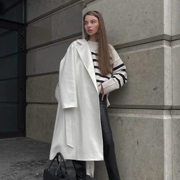 A woman strolls through SoHo in a beige trench coat layered over a striped knit and high-waisted denim. The outfit blends Parisian charm with NYC street style.